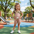 Young girl in a pink floral dress standing on a playground with colorful equipment and trees in the background.