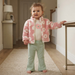 A toddler stands barefoot in a hallway wearing a pink fuzzy zip-up jacket with white flower patterns, a cream top with a pink flower applique, and light sage green pants.