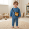 A toddler stands barefoot on a woven rug in a bright nursery, wearing a blue sweatshirt and matching pants with a small yellow lion-face crossbody bag.