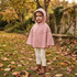 A young child wearing a pink hooded cape with ruffle trim and buttons, cream pants, and brown boots, standing outdoors on grass covered with fallen leaves in a garden setting.