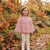 A young girl stands outdoors in a garden wearing a dusty pink knit cape cardigan with a ruffled yoke and button front, paired with cream leggings. The background features autumn foliage, fallen leaves, and pumpkins.