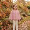 A young girl stands outdoors in a garden wearing a dusty pink knit cape cardigan with a ruffled yoke and button front, paired with cream leggings. The background features autumn foliage, fallen leaves, and pumpkins.