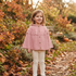 A young girl stands outdoors in a garden wearing a dusty pink knit cape cardigan with a ruffled yoke and button front, paired with cream leggings. The background features autumn foliage, fallen leaves, and pumpkins.