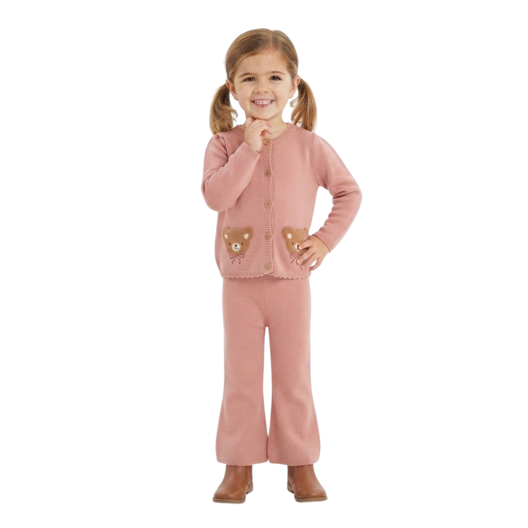 Young girl smiling with pigtails, wearing a dusty pink knit cardigan featuring two embroidered teddy bear pockets and matching flared trousers against a plain white background.
