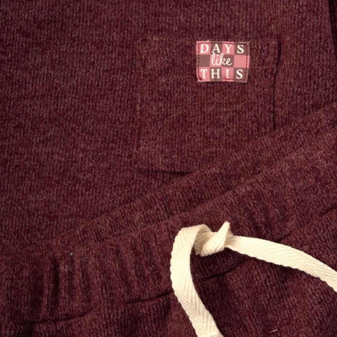 Close-up of a ribbed knit garment in deep burgundy with a cream drawstring and a small chest patch reading 'DAYS like THIS' in pink, burgundy, and dark gray.