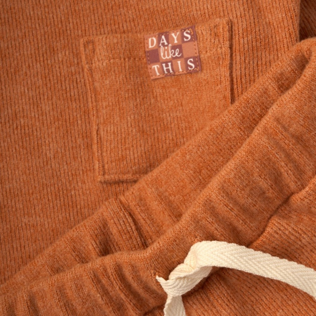 Close-up of an orange-brown ribbed knit garment with a front pocket and a sewn label that reads “DAYS like THIS.” A cream drawstring is visible at the bottom right.