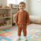 A toddler stands on a colorful alphabet play mat in a bright children's room, wearing a matching rust-orange knit long-sleeve top and jogger pants with white sneakers.