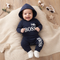 A baby lying on a beige knit blanket wearing a navy blue hooded sweatshirt and matching pants with white 'Little BOSS' text, styled with cotton stems, sunglasses, and star decorations on a wood floor.