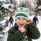 Close-up of a young boy smiling happily outdoors in the snow, wearing a white and green Minecraft bobble hat, a green coat, and a pixel-patterned scarf.