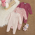 Three infant footed sleeper jumpsuits in soft corduroy fabric are arranged on a woven beige rug with a white faux-fur surface and a small plush toy. The set includes a light pink and white striped sleeper, a pink patterned sleeper, and a dusty rose/pink patterned sleeper.