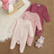 Three infant footed sleeper jumpsuits in soft corduroy fabric are arranged on a woven beige rug with a white faux-fur surface and a small plush toy. The set includes a light pink and white striped sleeper, a pink patterned sleeper, and a dusty rose/pink patterned sleeper.