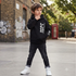 A young boy wearing a black hoodie with white graphic text, black jeans, and white sneakers, standing in an outdoor urban setting with a brick wall, graffiti, and a red telephone booth in the background.