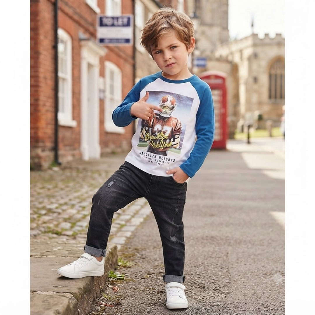 A young boy wearing a blue and white raglan t-shirt with a graphic print, paired with dark jeans and white sneakers, posing outdoors on a street with a red telephone booth and brick buildings in the background.