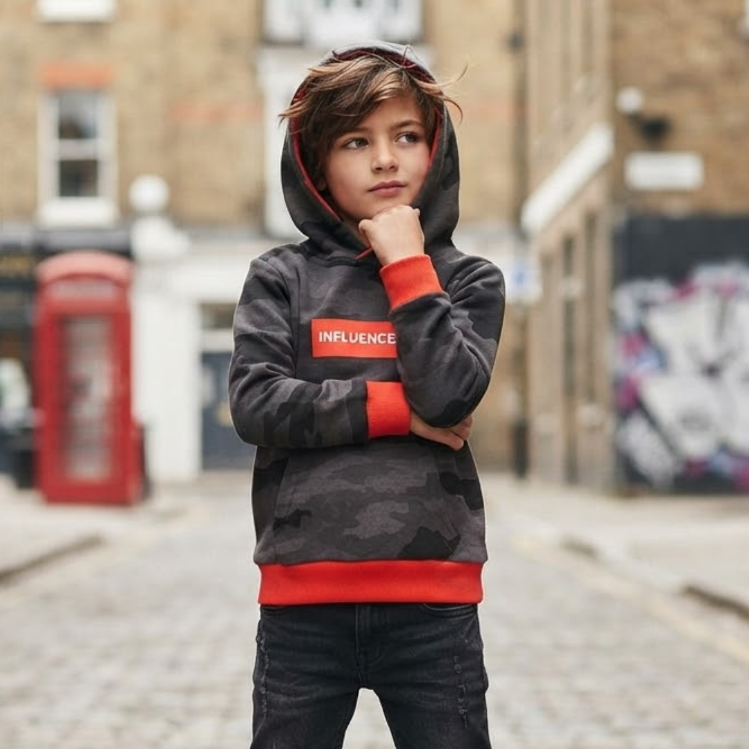A child wearing a dark gray and black camouflage hoodie with red accents and the word 'INFLUENCE' on the chest, standing on a cobblestone street with a blurred background.
