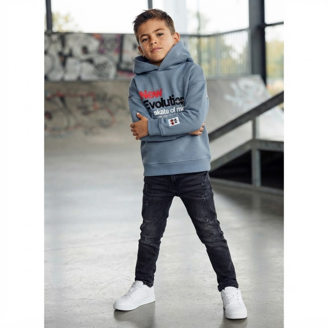 A young boy wearing a light blue hoodie with 'New Evolution style of life' printed on the front, paired with dark jeans and white sneakers, standing in an indoor skatepark.