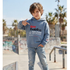 A young boy wearing a light blue hoodie with 'Now Evolution' printed on the front and light-wash jeans, standing in an outdoor skatepark setting.