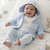 A smiling baby wearing a light blue knit hooded cardigan and matching pants over a white t-shirt with 'Prince' embroidered on the chest, lying on a white quilted bed.