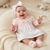 A smiling baby girl wearing a light pink long-sleeve dress with a white patterned skirt, a white bow headband, and pink booties, sitting on a cream knit lounger.
