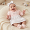 A smiling baby girl wearing a light pink long-sleeve dress with a white patterned skirt, a white bow headband, and pink booties, sitting on a cream knit lounger.