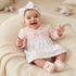 A smiling baby girl wearing a light pink long-sleeve dress with a white patterned skirt, a white bow headband, and pink booties, sitting on a cream knit lounger.