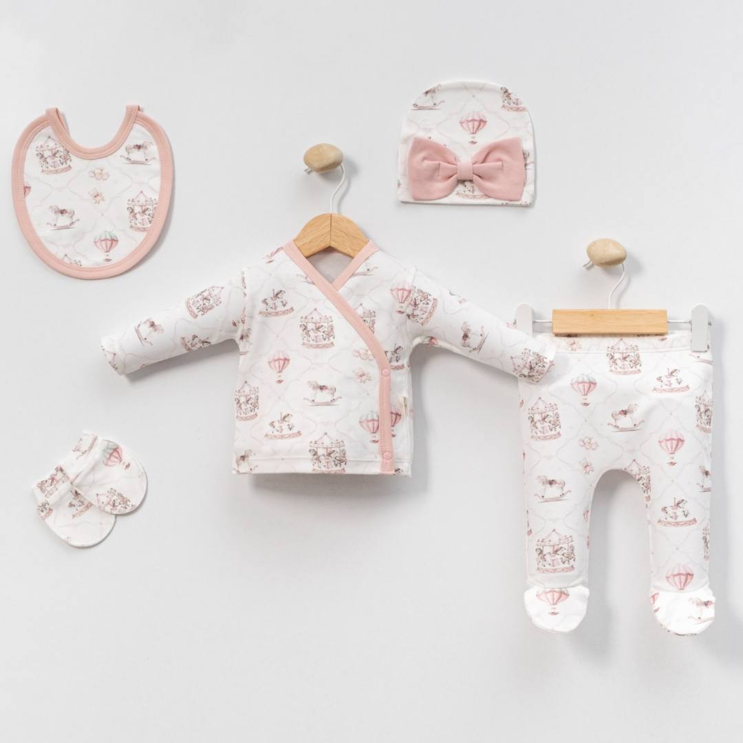A baby clothing set displayed on a light gray background, including a long-sleeve wrap jacket on a wooden hanger, matching footed pants on a white hanger, a bib, a bow headband, and a small bootie. The garments are white with a pink and beige hot air balloon and teddy bear pattern.