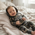 A baby lying on a cozy beige knit blanket, wearing a camouflage hooded one-piece outfit with 'DADDY'S BOY' printed across the chest.