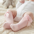A close-up of a baby's legs wearing light pink knit booties with large bow details, lying on a cream cable-knit blanket.