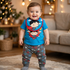 Smiling baby wearing a Superman outfit in a warmly lit, festively decorated room.