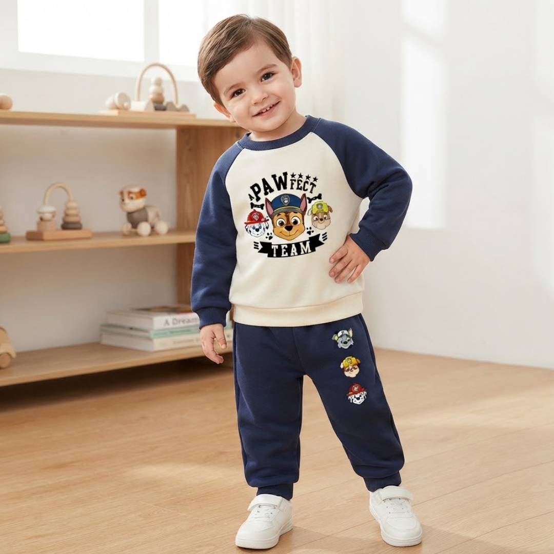 A toddler boy wearing a navy blue and cream 2-piece pajama set with a Paw Patrol graphic on the sweatshirt and matching character prints on the pants, standing in a brightly lit child's room.