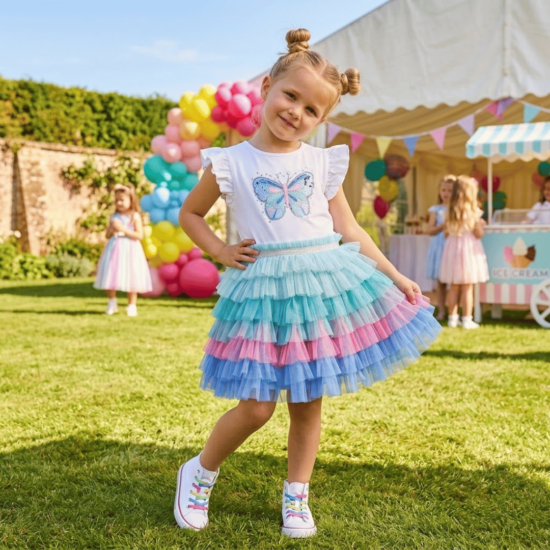 Young girl in a colorful skirt with a butterfly design standing outdoors with balloons and a cart in the background.