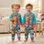 Two young children smiling while standing side-by-side in matching turquoise and pink Paw Patrol fleece onesies in a cozy living room.
