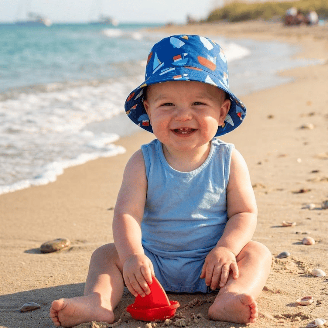 Child wearing a blue sun hat with boat pattern on a sandy beach.