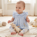Baby in a light blue dress with bunny design sitting on a soft surface holding a plush toy.