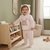 A baby wearing a light pink two-piece fleece outfit with a bunny graphic on the chest and a bear-ear hood, standing in a cozy nursery next to a wooden toy organizer.
