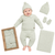 A baby wearing a light sage green knit three-piece outfit with a matching beanie and mittens, shown alongside the outfit packaging, a folded blanket, and additional accessories on a white background.
