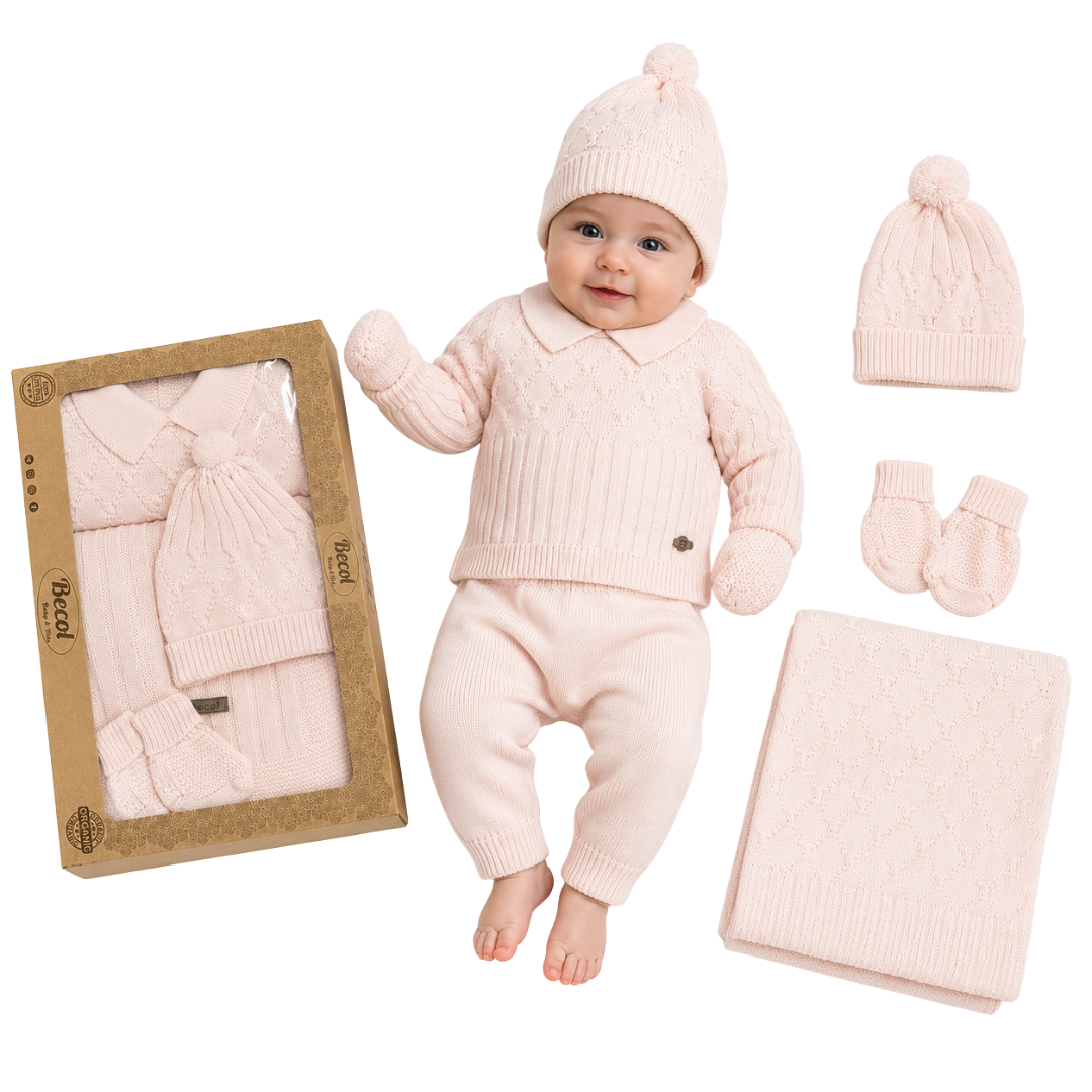 A baby wearing a light pink knit three-piece outfit with a matching beanie, mittens, and blanket, displayed next to its kraft paper packaging box and an additional matching set on a white background.