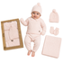 A baby wearing a light pink knit three-piece outfit with a matching beanie, mittens, and blanket, displayed next to its kraft paper packaging box and an additional matching set on a white background.