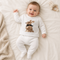 A baby lying on a white blanket wearing a white long-sleeve top and matching white pants, with a brown rabbit graphic on the chest. A beige knit blanket is on the left, and a small stuffed bunny toy is on the upper right.