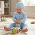 A baby wearing a light blue textured knit one-piece outfit with a matching hat sits on a patterned rug, smiling while playing with a wooden shape-sorting toy. The room is softly lit with natural light from a nearby window.