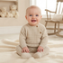 A smiling baby sitting on a cream blanket in a softly lit nursery, wearing a beige ribbed knit footed romper with brown buttons.