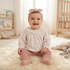 A smiling baby girl sitting on a fluffy cream rug in a bright nursery, wearing a cream long-sleeve lace top, dusty pink knit tights, and a dusty pink bow headband.