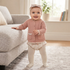A baby standing indoors near a sofa, wearing a dusty pink textured cardigan with front pockets, cream tights, a floral diaper cover, and a matching bow headband.