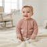 A smiling baby sitting on a cream blanket in a bright nursery, wearing a dusty pink textured cardigan with white lace trim, matching pink knit tights, and a pink bow headband.