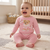 A baby sitting on a cream rug in a nursery, wearing a pink knit two-piece outfit with a teddy bear graphic on the top and matching pink pants.