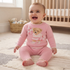 A baby sitting on a cream rug in a nursery, wearing a pink knit two-piece outfit with a teddy bear graphic on the top and matching pink pants.
