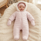 A baby wearing a light pink quilted hooded footless sleeper with matching mittens and a knit hat, lying on a fluffy cream blanket in a softly lit nursery.