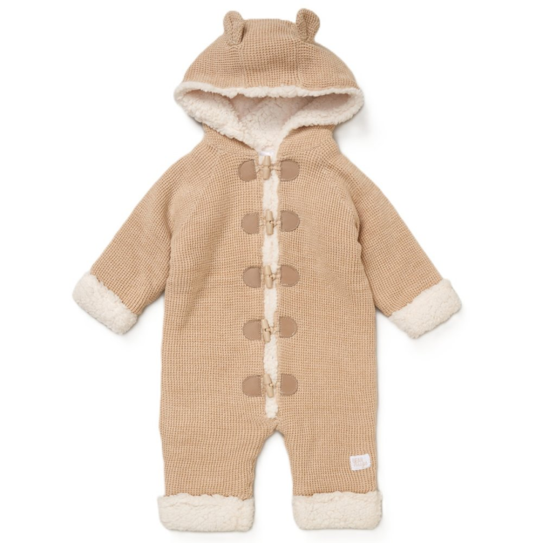A beige ribbed knit hooded baby jumpsuit with cream sherpa lining, featuring toggle closures down the front, a hood with bear ears, and matching cream sherpa trim at the cuffs and ankles.