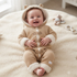 A baby wearing a cozy knit hooded one-piece outfit in beige with cream faux-fur trim, sitting on a soft cream blanket in a bright room. The outfit has bear-ear details on the hood and front button closures.