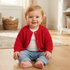 A baby sitting on a light wood floor wearing a bright red knit cardigan over a white shirt and blue jeans.