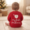 A baby sitting on a wooden floor, viewed from behind, wearing a red knit sweater with a white Santa face and the words 'my first christmas' written in white script.